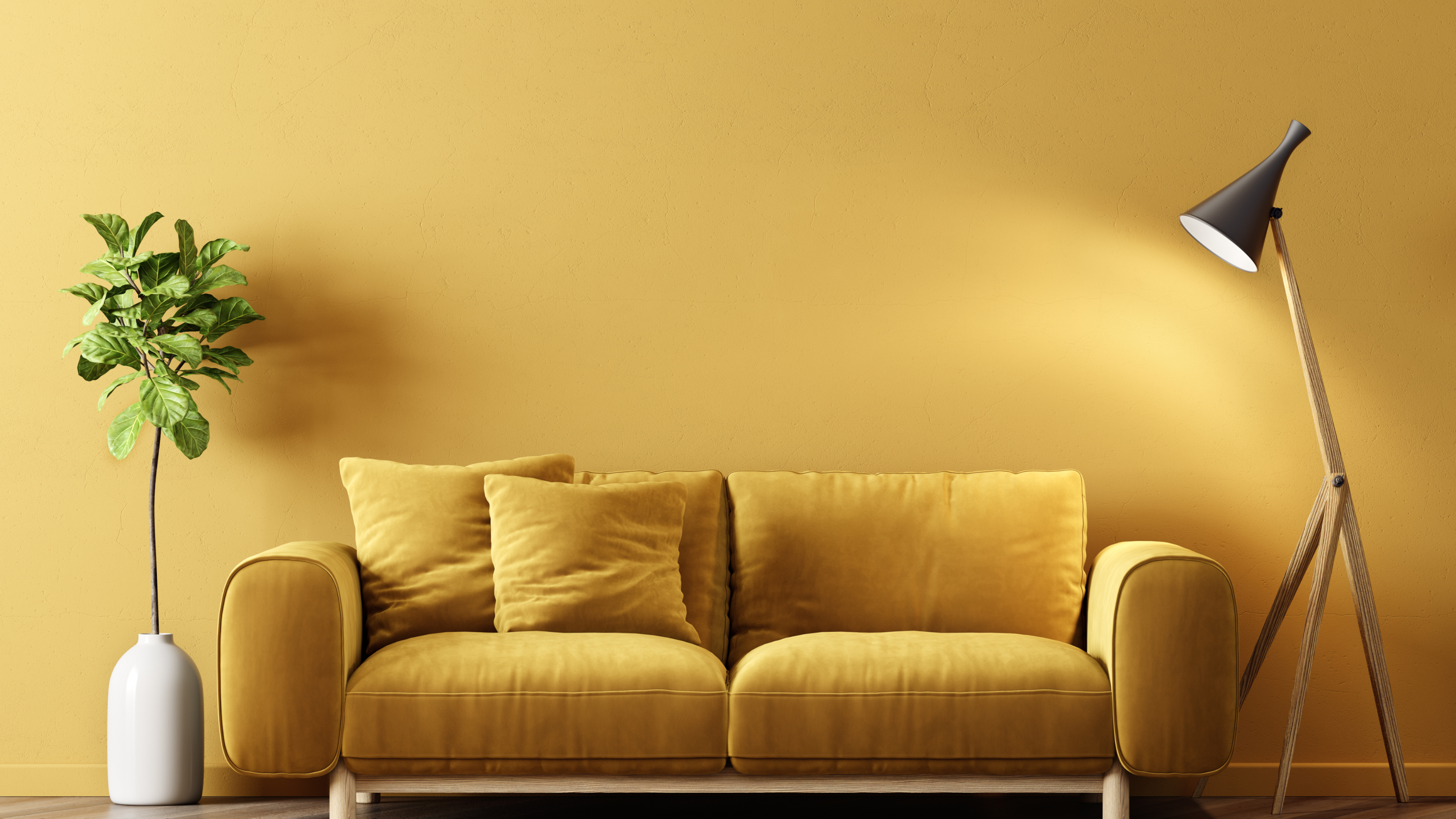 A cozy yellow velvet sofa with cushions, accompanied by a tall plant in a white vase and a stylish wooden lamp against a matching wall.
